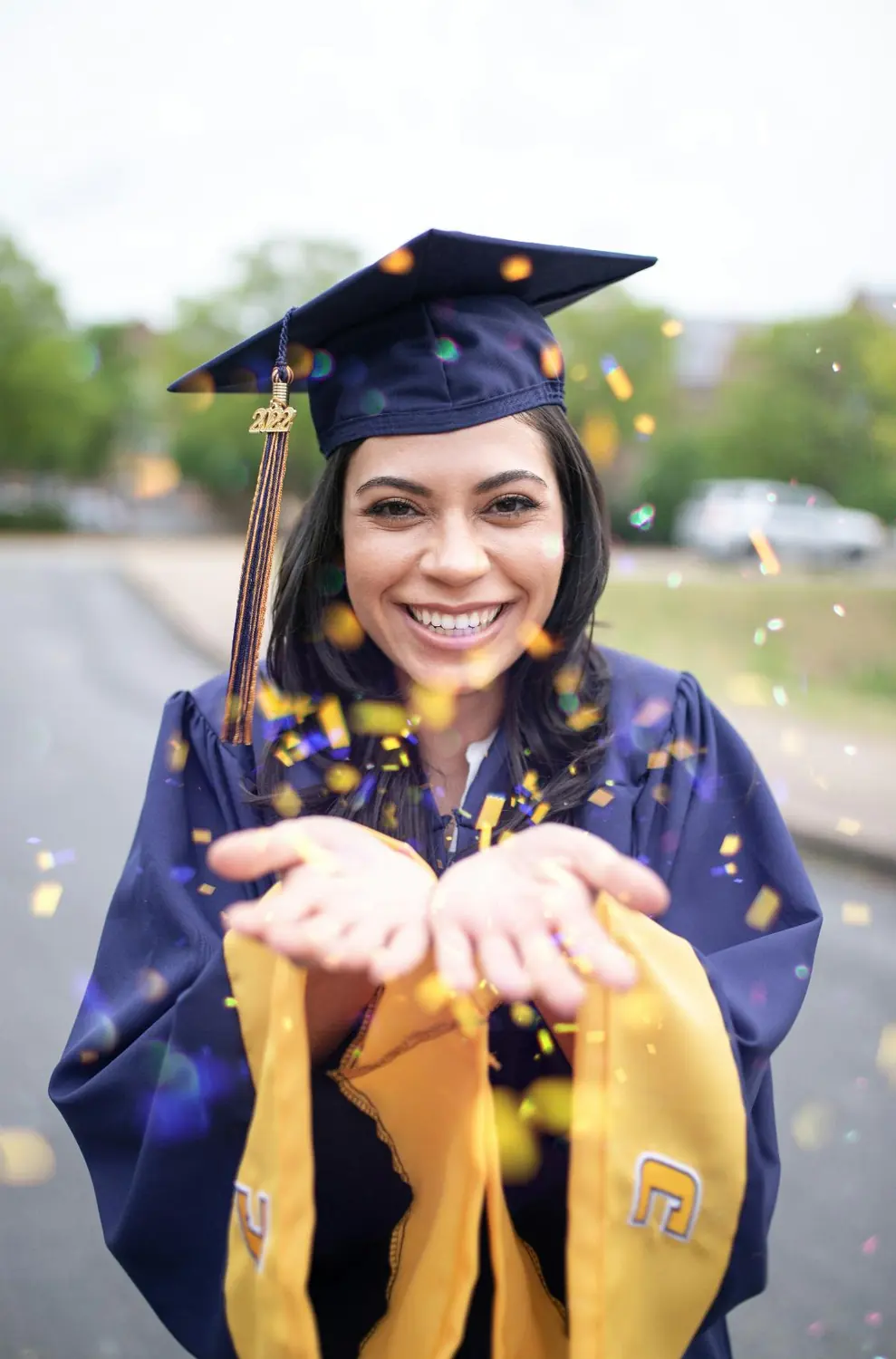 Graduate in navy cap and gown throwing gold confetti toward the camera
