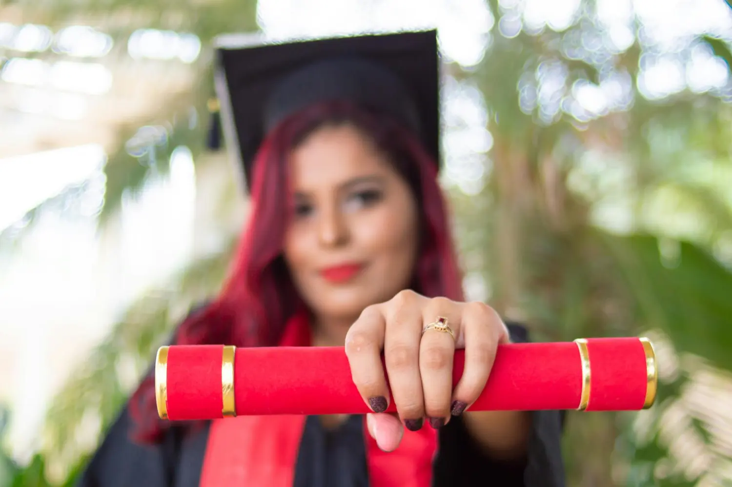 Graduate holding a rolled red diploma toward the camera