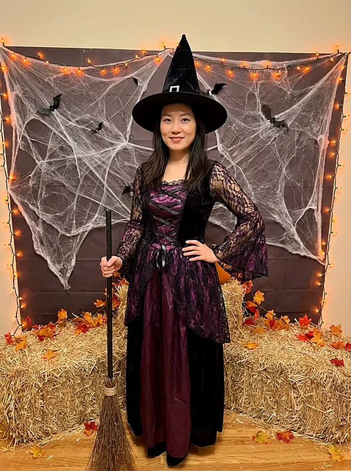 Smiling woman in a black witch hat and lace dress holding a broom in a cobweb-decorated room, generated by the Halloween photo pack